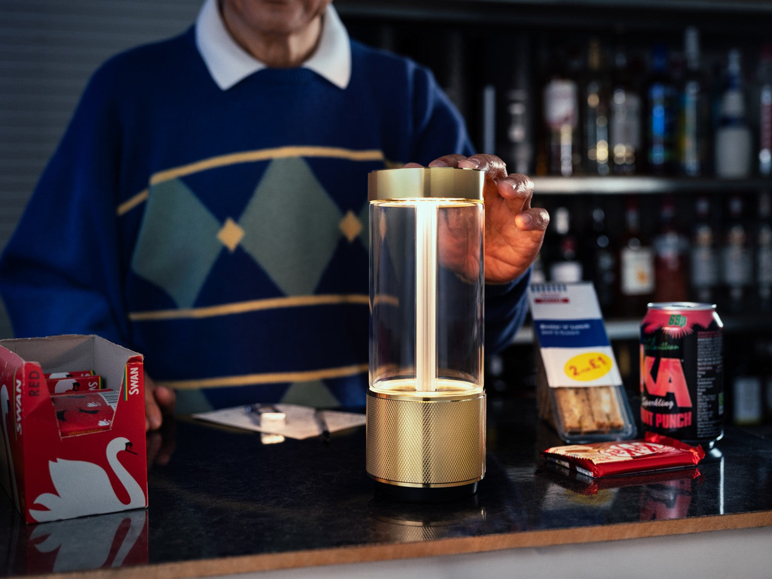 Person holding a gold and clear cylindrical lamp with a bar setting in the background.